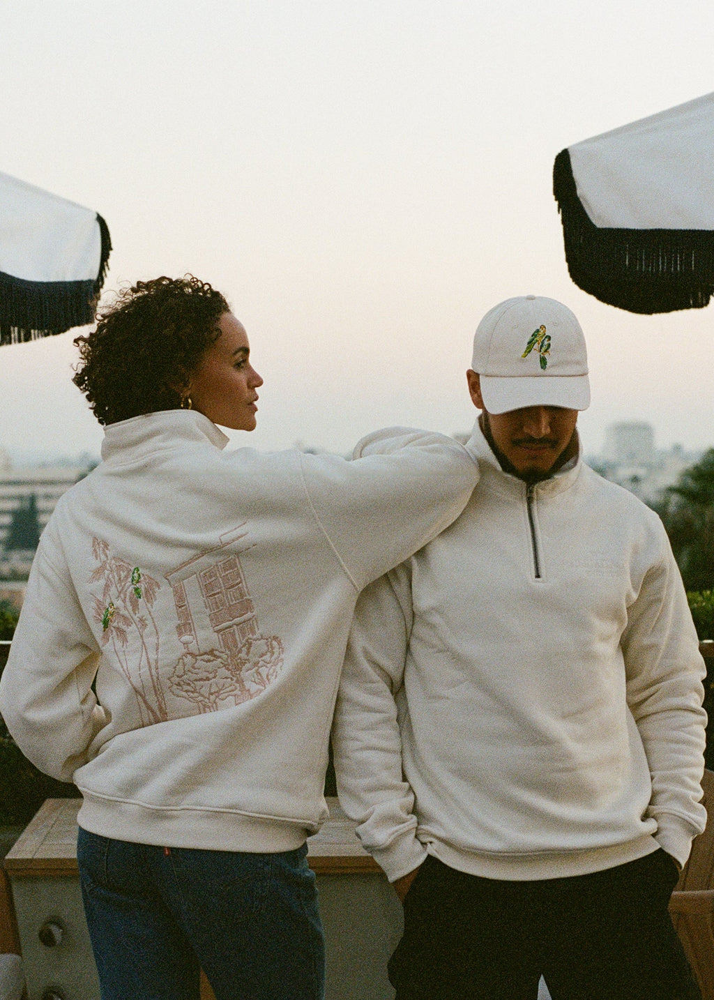 Two people wearing the L'Ermitage Beverly Hills x Dinner Service NY limited merch collection standing on a rooftop with cityscape in the background.